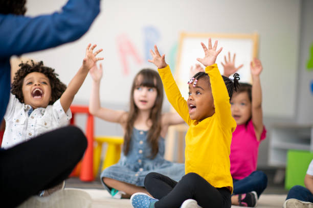 A multi-ethnic group of preschool students is sitting with their legs crossed on the floor in their classroom. The mixed-race female teacher is sitting on the floor facing the children. The happy kids are smiling and following the teacher's instructions. They have their arms raised in the air.
